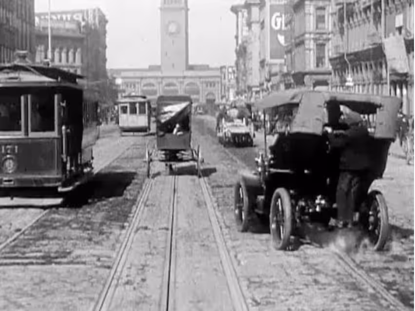 San Francisco Market Street 1906 Film Still. Greyscale image. Half a tramcar is visible left of 3:4 frame. We are looking down a street. Large buldings are in the background to the right and left. Ahead is a building with a substantial clock tower. On the left is an early motor car with a fabric roof. In the centre a horse drawn cart.