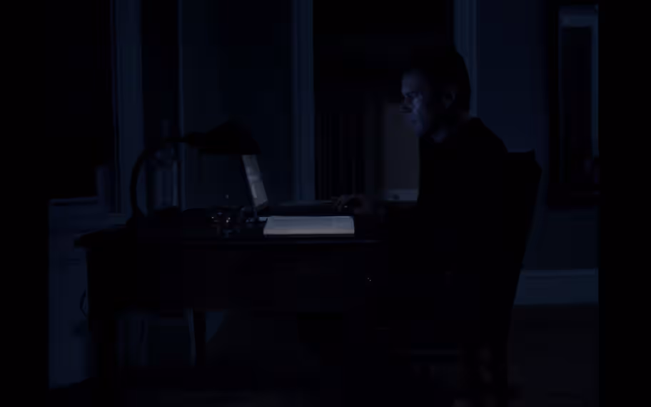 Film Still from First Reformed, 2017. A mid-frame of a white man with short dark hair in profile sat in a dark room, barely lit by the glow of his computer screen