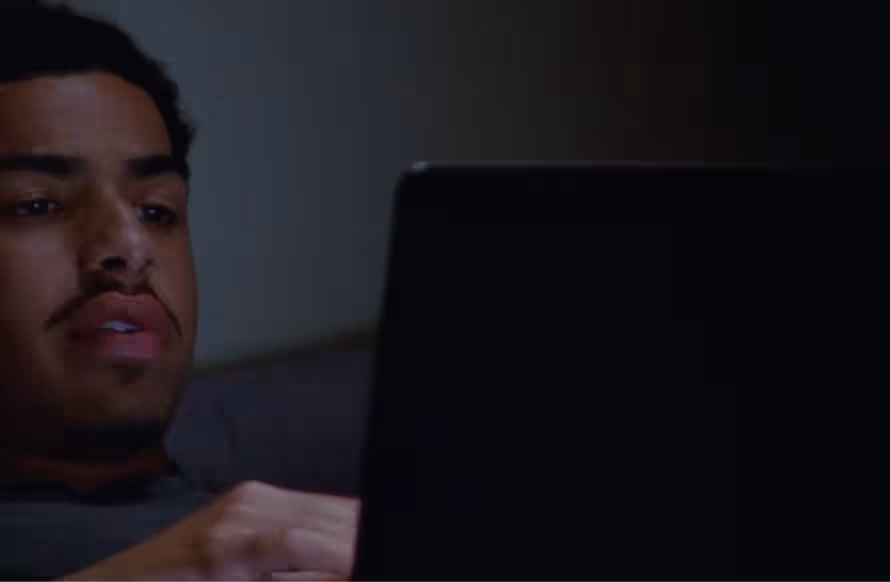 Film Still from How to Blow Up a Pipeline, 2022. A close up of a young Black man with short thick black hair and a light moustache on a sofa on a laptop. His hand and face are lit by the screen.