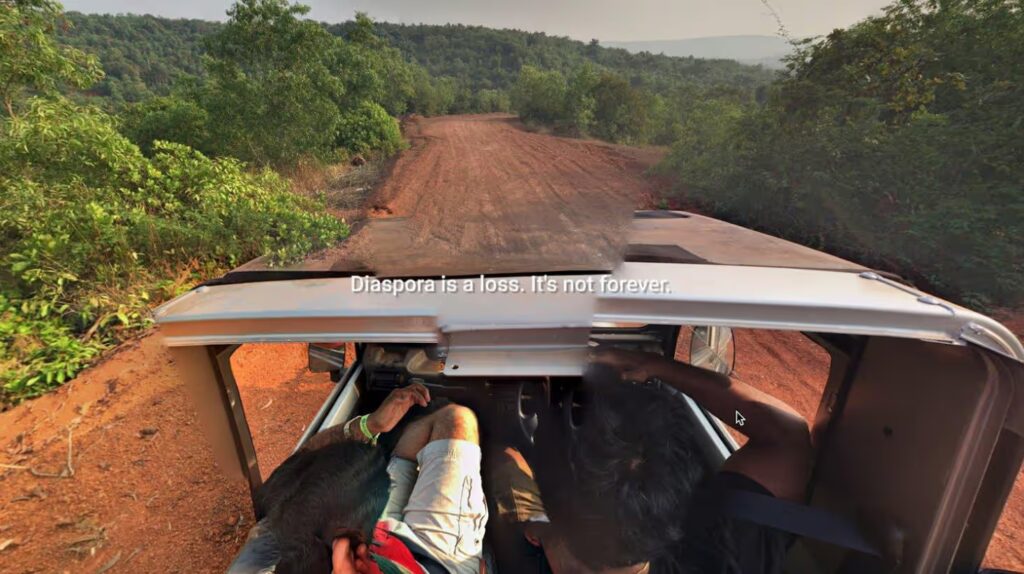 Film still from 'At Home but not at Home', 2019. We see a fragmented 'Google Maps Streetview' style image showing an open topped vehicle and the backs of its human occupantsof an orange dirt track through a heavily vegetated, perhaps tropical landscape. Overlaid across the centre of the image are the words "Diaspora is a loss. It's not forever."