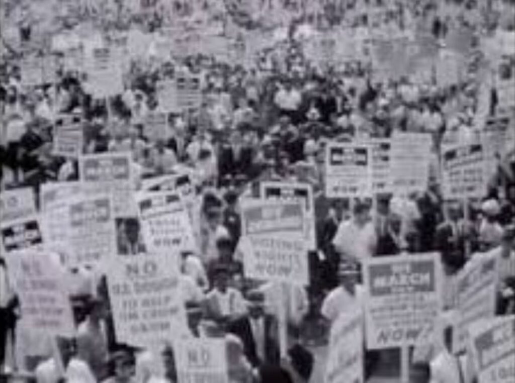 Film still from 'Love is the Message, but the Message is Death', 2017, showing an archive greyscale image of a US Civil Rights protest demanding voting rights NOW. Many people are in the frame, many carrying placards.