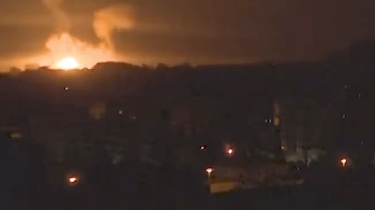 Film Still from Happiness 2025. We are overlooking a darkened city. A few streetlights and indistinct buildings are visible. A very bright light is visible on the horizon to the left, along with what might be smoke. It is either the sun rising or an explosion. It looks like it could be from a war, perhaps Gaza.